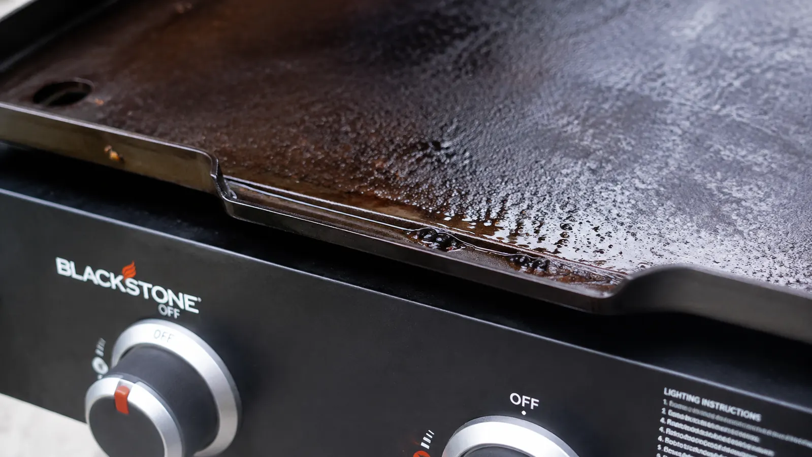 Grease pooled at the edge of a cast iron griddle on a camp stove, dripping toward the burner control knobs
