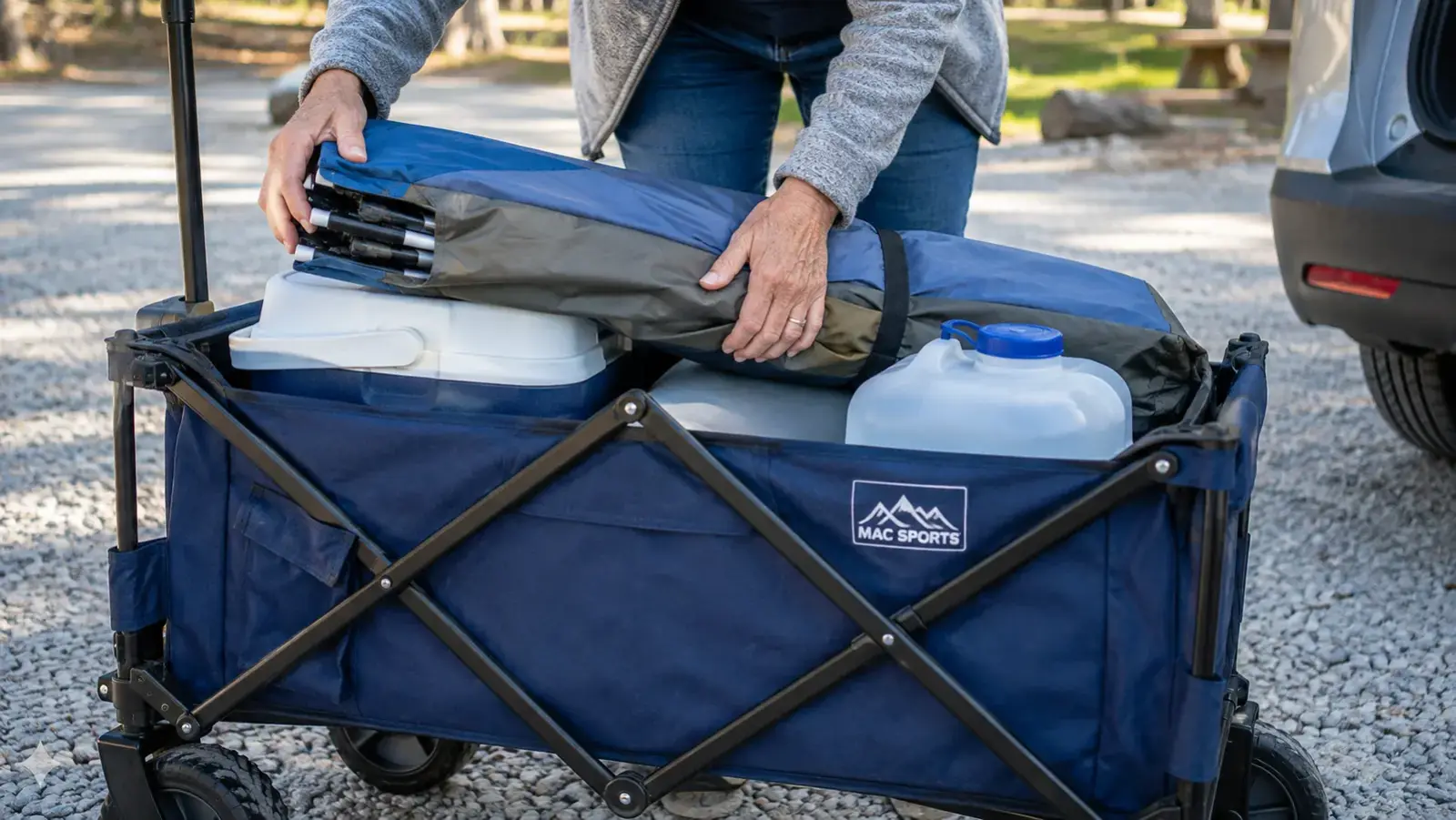 Close-up of hands loading a cooler, folded tent, and water jug into a collapsible wagon at a gravel parking pad